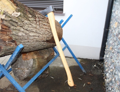 Siekiera leśna 1,6 kg Krumpholz z drewnianą rączką, leżąca na pniu w poziomie, z metalowym ostrzem z przodu. Tło proste, budynek w jasnym kolorze. Rączka długa, gładka, jasna, w kolorze naturalnego drewna.