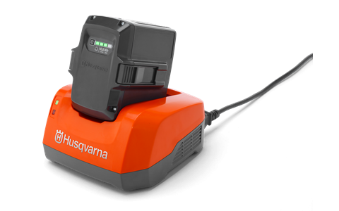 Ładowarka do akumulatorów o pomarańczowej podstawie z szarymi detalami, z włożonym akumulatorem. Widok z przodu, z widocznymi wskaźnikami naładowania. Produkt leży na prostym, ciemnym tle, podłączony do zasilania.