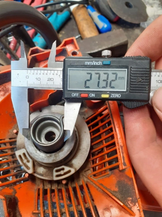 Sprężyna rozrusznika pilarki Husqvarna 543X/543XPG, widok z góry, na płaskiej powierzchni. Metalowe elementy, białe oznaczenia, długość 27,32 mm mierzone suwmiarką. Tło z narzędziami warsztatowymi.
