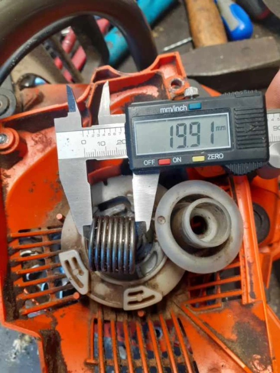 Sprężyna rozrusznika 19,91 mm w czerwonej obudowie pilarki Husqvarna, z widocznymi mocowaniami i oznaczeniami, widok z boku na elementy mechaniczne na tle metalowych narzędzi.