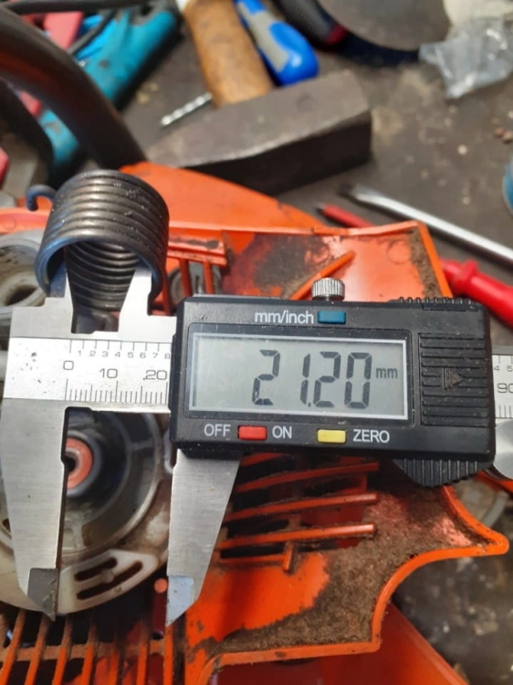Sprężyna rozrusznika do pilarki 543X/543XPG Husqvarna, widoczna z boku. W tle pomiar 21,20 mm na elektronicznym mikrometrze. Element metalowy, sprężysty, o okrągłym kształcie. Otoczenie robocze, narzędzia oraz części mechaniczne w tle.