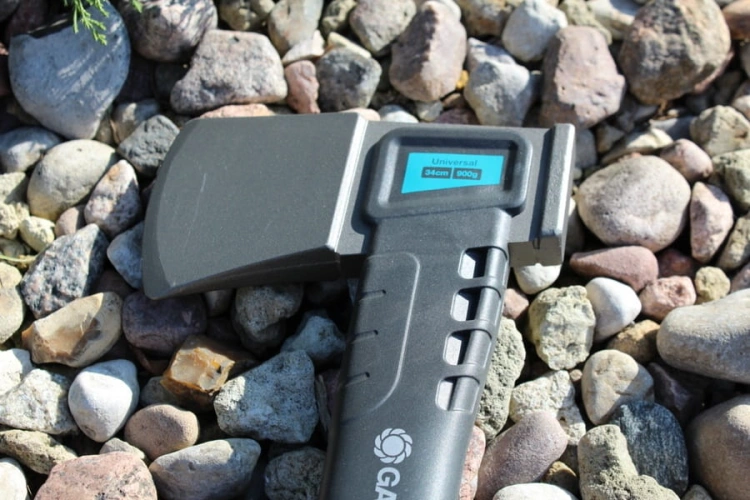 Siekiera uniwersalna 900B GARDENA z prostokątną głowicą, czarnym teksturowanym uchwytem i niebieskim wyświetlaczem, 34 cm, 900 g, leżąca na kamieniach, widok z boku pokazuje uchwyt i ostrze.