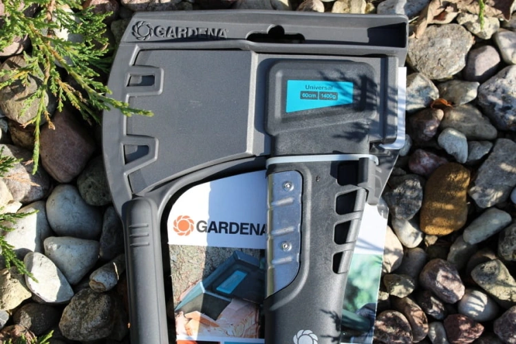 Siekiera uniwersalna GARDENA 1400A w kolorze czarnym z szaro-niebieskimi detalami. Ujęcie frontalne, stojąca na tle różnych kamieni. Widać napis Universal oraz wymiary: 60 cm, 1400 g. Rączka z wypustkami dla lepszego uchwytu.