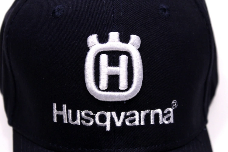 Granatowa czapka z daszkiem Husqvarna, pokazana z bliska, z widocznym białym logo oraz napisem Husqvarna. Czapka ma klasyczny kształt, wykonana z materiału o gładkiej teksturze. Tło białe, co podkreśla detale.