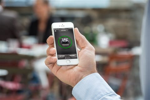 Smartfon w dłoni z aplikacją do zdalnego sterowania robotem koszącym Husqvarna Automower® 450X. Ekran wyświetla ikonki 'STOP' i 'PAUSE'. Tło przedstawia rozmyte krzesła i stoliki w ogrodzie.
