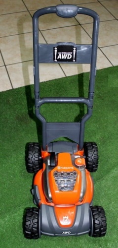 Zabawka dla dzieci Kosiarka AWD Husqvarna w kolorze pomarańczowym i szarym, widoczna z przodu. Posiada plastikowy uchwyt, duże, czarne koła oraz teksturę imitującą silnik. Tło stanowi sztuczna trawa.