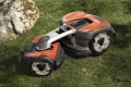 Automower® 535 AWD robot koszący Husqvarna w pozycji stojącej na trawie, z boku. Posiada czerwono-czarną obudowę z metalowym elementem z przodu. Widoczne duże koła z bieżnikiem i logo Husqvarna na górze. W tle kamień naturalny.