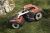 Automower® 535 AWD robot koszący Husqvarna w pozycji stojącej na trawie, z boku. Posiada czerwono-czarną obudowę z metalowym elementem z przodu. Widoczne duże koła z bieżnikiem i logo Husqvarna na górze. W tle kamień naturalny.