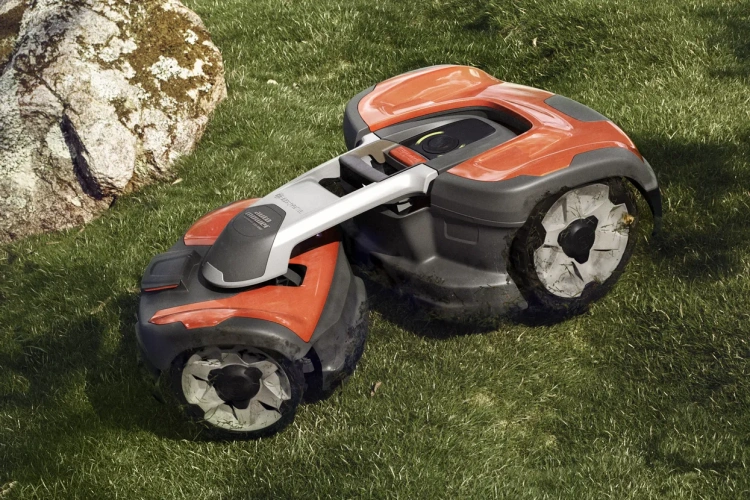 Automower® 535 AWD robot koszący Husqvarna w pozycji stojącej na trawie, z boku. Posiada czerwono-czarną obudowę z metalowym elementem z przodu. Widoczne duże koła z bieżnikiem i logo Husqvarna na górze. W tle kamień naturalny.