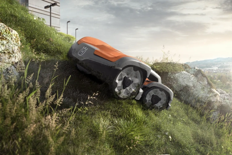 Robot koszący Automower® 535 AWD Husqvarna w kolorze szaro-pomarańczowym, widoczny z boku na stromym, zielonym terenie, z widocznymi kołami i logo producenta. Tło przedstawia naturalne otoczenie z kamieniami i trawą.