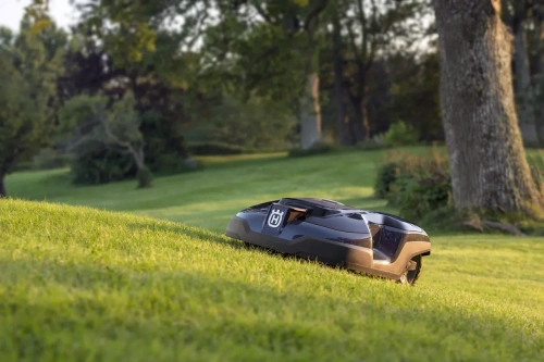 Automower® 315 Mark II to robot koszący z czarną obudową i logo Husqvarna, widoczny w ujęciu z boku na pochyłym terenie. Produkt znajduje się na świeżo skoszonej trawie, w tle widoczne drzewa i bujna zieleń.