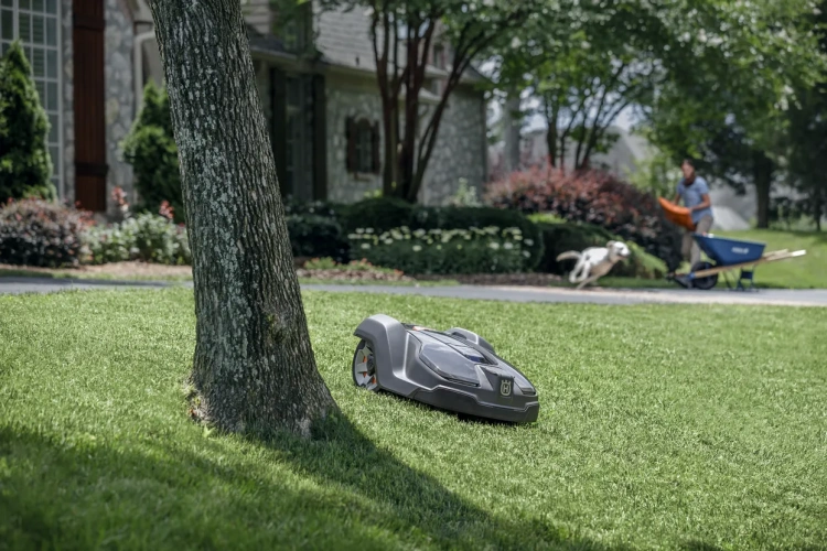 Automower® 430X szary robot koszący Husqvarna na trawie, z logo. Widok z boku, pokazuje zaokrąglony kształt i boczne krawędzie. W tle zieleń i osoba z wózkiem ogrodowym.