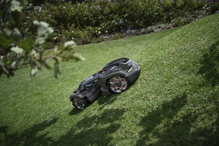 Robot koszący Husqvarna Automower® 435X AWD w kolorze ciemnoszarym, z widocznymi detalami na kołach. Pozycjonowany na nachylonym trawniku, z widokiem z boku. Otoczenie z zieloną trawą i lekkim cieniem roślin.