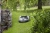Automower® 450X robot koszący w kolorze czarnym, z widocznym logo Husqvarna, leżący na trawie w ogrodzie. Ujęcie z boku pokazuje jego aerodynamiczny kształt oraz system czujników otaczających. W tle drzewa i kamienia tworzą naturalne otoczenie.
