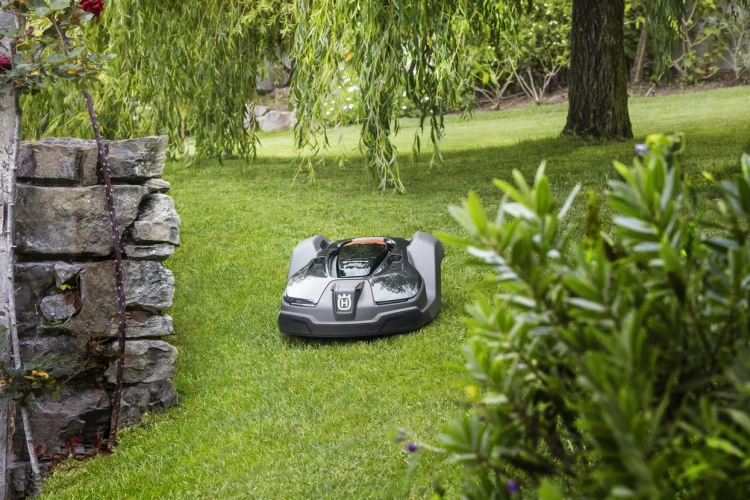 Automower® 450X robot koszący w kolorze czarnym, z widocznym logo Husqvarna, leżący na trawie w ogrodzie. Ujęcie z boku pokazuje jego aerodynamiczny kształt oraz system czujników otaczających. W tle drzewa i kamienia tworzą naturalne otoczenie.