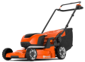 Kosiarka akumulatorowa LC247i Husqvarna w pomarańczowym kolorze z czarnymi elementami. Ujęcie z boku, pokazujące twardą obudowę i zbiornik na trawę. Widoczna regulowana rączka oraz duże, gumowe koła. Produkt stoi na podłożu.