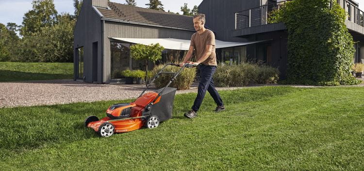 Kosiarka akumulatorowa LC142iS Husqvarna w kolorze pomarańczowym, używana w ogrodzie. Ujęcie z boku pokazuje mężczyznę prowadzącego kosiarkę po trawie. Tło wypełnione roślinnością i nowoczesnym domem. Widoczny tylny zbiornik na trawę.