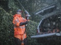 Spodnie przeciwdeszczowe Vent Technical Husqvarna w intensywnym pomarańczowym kolorze, z kontrastowymi, odblaskowymi paskami. Model rozłożony na mężczyźnie, trzymającym narzędzie, w deszczowej scenerii. Tło z otwartym bagażnikiem pojazdu.