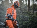 Kurtka przeciwdeszczowa Husqvarna w kolorze jaskrawego pomarańczu, widoczna na modelu w deszczu. Odporny materiał z odblaskowymi elementami. Ujęcie z boku, podkreślające ergonomiczne detale oraz funkcjonalność w trudnych warunkach.