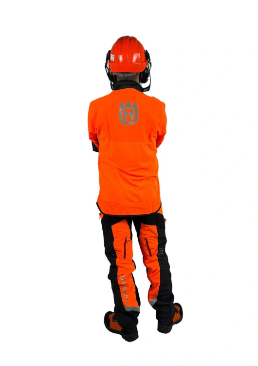 Kask leśny Classic Husqvarna w kolorze pomarańczowym z ciemnymi akcentami. Ujęcie z tyłu, model w pozycji stojącej. Widoczne logo firmy na plecach oraz elementy odblaskowe na nogawkach. Tło jednolite, białe.