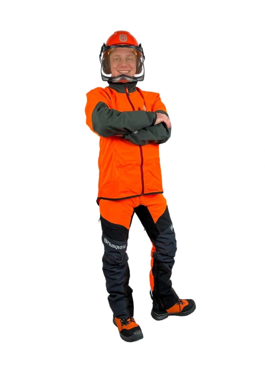 Kask leśny Classic Husqvarna w pomarańczowym kolorze z przezroczystą osłoną, trzymany przez mężczyznę w odzieży roboczej pomarańczowo-czarnej z logo Husqvarna, zapewniający bezpieczeństwo podczas pracy w lesie.