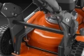 Kosiarka spalinowa Husqvarna LB 251S, widoczna z boku. Na zdjęciu widać pomarańczową obudowę silnika, czarną kierownicę z regulacją, metalowe elementy mocujące oraz czarne koła. Tło jest jednolite, co podkreśla detale produktu.