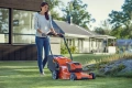 Kosiarka akumulatorowa LC353iVX Husqvarna w kolorze pomarańczowym. Widoczna z boku, użytkownik prowadzi ją po zielonym trawniku. Na zdjęciu teren z zabudową w tle, kosiarka z przodu ma duży, czarny kosz na trawę i wyraźne koła.