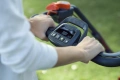 Kosiarka akumulatorowa Husqvarna LC353iVX trzymana w dłoniach, zbliżenie na uchwyt i panel sterujący. Wyraźne przyciski oraz wskaźnik poziomu naładowania, czarny uchwyt z dodatkami w kolorze pomarańczowym. Tło zielone, trawiaste.