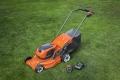 Pomarańczowa kosiarka akumulatorowa Husqvarna LC347iVX na trawie, z czarnymi akcentami. Widoczny akumulator, ładowarka, przednia listwa tnąca i pojemnik na trawę, ujęcie z boku.