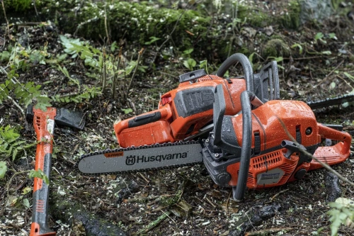 Pilarka Husqvarna 550XP Mark II w kolorze pomarańczowym, leżąca na ziemi z mokrymi kroplami. Widoczny uchwyt, ząbkowany łańcuch z oznaczeniem Husqvarna, blisko leżąca siekiera. Tło to leśna ziemia pokryta liśćmi i gałęziami.