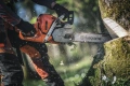 Pilarka Husqvarna 572 XP® w pomarańczowym kolorze, z boku pokazany użytkownik tnący drzewo ostrą metalową prowadnicą. Leśne tło z kawałkami drewna w powietrzu.