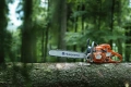 Pomarańczowa pilarka Husqvarna 562 XP®G na pniu z metalową prowadnicą, widoczny bok z logo i tłoczoną obudową. Ergonomiczne uchwyty, tło z zielonymi drzewami.