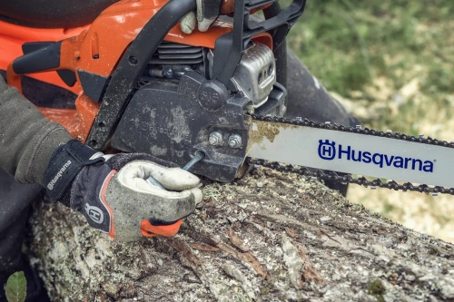 Pilarka Husqvarna 135 Mark II w zbliżeniu, z widocznym logo na łańcuchu. Narzędzie znajduje się w dłoni użytkownika, który przytrzymuje klucz przy mocowaniu obok silnika. Tło leśne, widoczne pęknięcia kory na pniu.