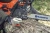 Pilarka Husqvarna 135 Mark II w zbliżeniu, z widocznym logo na łańcuchu. Narzędzie znajduje się w dłoni użytkownika, który przytrzymuje klucz przy mocowaniu obok silnika. Tło leśne, widoczne pęknięcia kory na pniu.