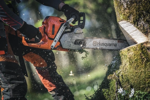 Pomarańczowa pilarka Husqvarna 572 XP® G z metalową prowadnicą, używana do cięcia drewna. Ujęcie z boku pokazuje rękojeść, logo na prowadnicy, kurz i odłamki drewna w naturalnym tle.