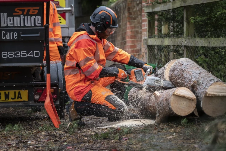 Pracownik w pomarańczowym stroju z odblaskami używa pilarki akumulatorowej Husqvarna 540i XP do cięcia grubych pni drzew. W tle drzewa i przyczepa, wióry drewna opadają podczas pracy.