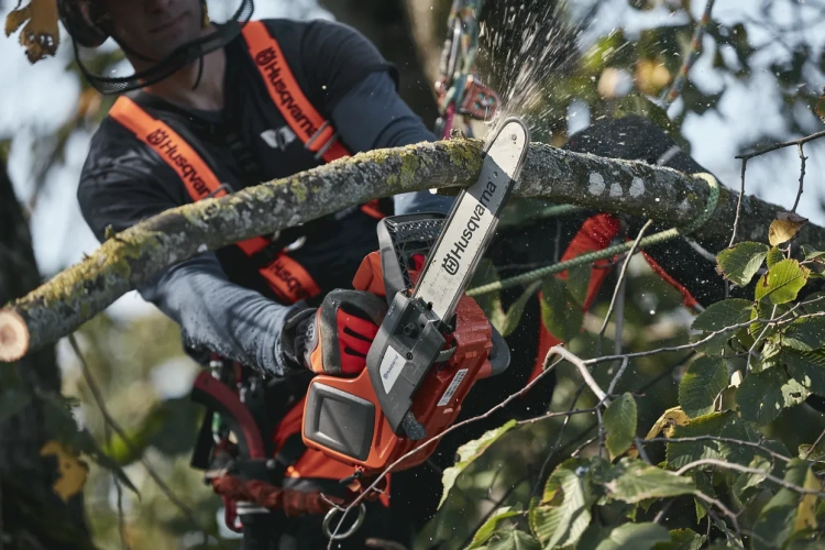 Pilarka akumulatorowa T535i XP Husqvarna trzymana w dłoni, zbliżenie na ostrze i korpus w kolorze pomarańczowym i czarnym. Urządzenie w trakcie cięcia gałęzi, otoczone zielonymi liśćmi. Widać konkretne detale, takie jak przyciski i uchwyt.