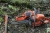 Pomarańczowa pilarka Husqvarna 545 Mark II na mokrej ziemi obok siekiery, z widocznym łańcuchem i logo na prowadnicy oraz ergonomiczną rączką na tle drewnianej posypki. Narzędzie wygląda na nowe.