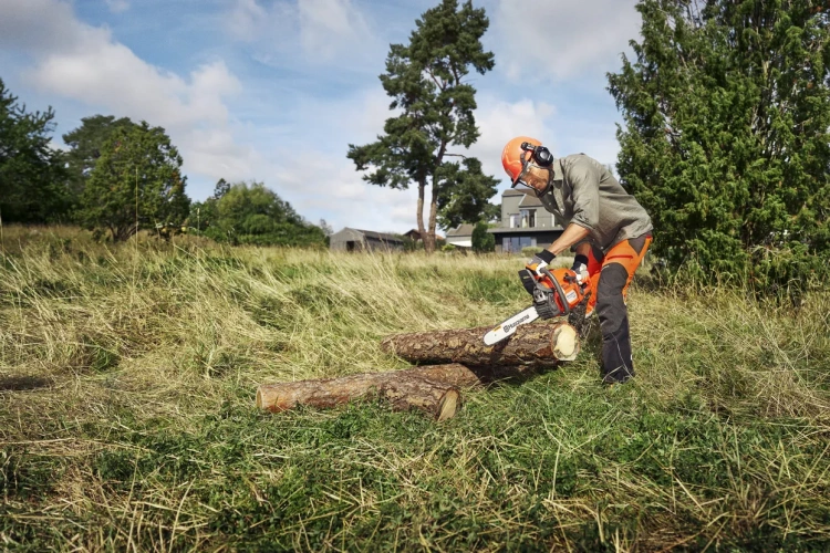 Mężczyzna w roboczym ubraniu tnie pilarką Husqvarna 435 II drewniane kłody na tle trawy i drzew. Pilarka stoi bokiem, z widocznym logo, lekka z pomarańczowymi akcentami.