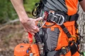 Pelarka Husqvarna T525, na zdjęciu przyczepiona do uprzęży, z widocznymi kolorami pomarańczowym i czarnym. Produkt prezentowany z boku, w dłoni trzymany klamra do mocowania, w otoczeniu naturalnym. W tle zarys drzew.