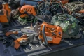 Pomarańczowa pilarka Husqvarna T525 z logo na obudowie na szarej powierzchni. W tle kask, rękawice i siatka z narzędziami, podkreślające kontekst pracy.