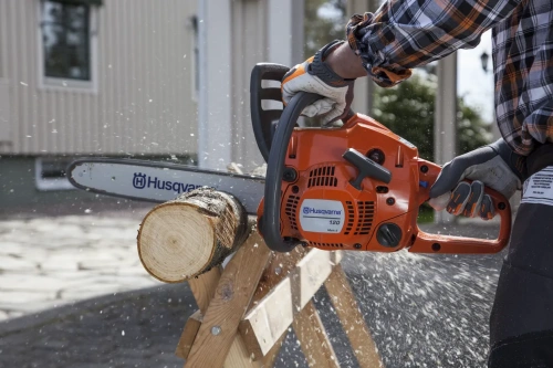 Pilarka Husqvarna 120 Mark II w pomarańczowej obudowie, trzymana w ręce przez osobę w rękawicach, zbliżenie na ostrze cięcia drewna, od czoła. W tle widoczna drewno w podporze. Wydobywające się wióry i krople wody.
