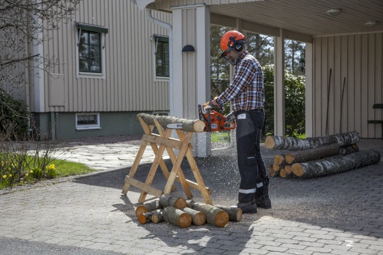 Mężczyzna w koszuli w kratę używa pilarki Husqvarna 120 Mark II do cięcia drewna na stojaku na tle jasnych ścian i stosów nieprzyciętego drewna z widocznymi okrągłymi końcami.