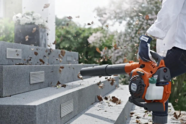 Dmuchawa-odkurzacz Husqvarna 125BVx w pomarańczowo-czarnym kolorze, używana stojąco. Użytkownik trzyma ją na schodach, liście lecą wokół. Matowa obudowa z widocznym logo Husqvarna.