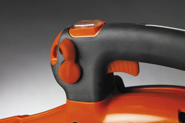 Zbliżenie na uchwyt dmuchawy Husqvarna 580BTS, w kolorze pomarańczowym i czarnym. Widoczne przyciski, w tym czerwony włącznik oraz regulacja mocy. Uchwyt ma ergonomiczną teksturę, co ułatwia trzymanie. Tło jest jednolite, ciemne.