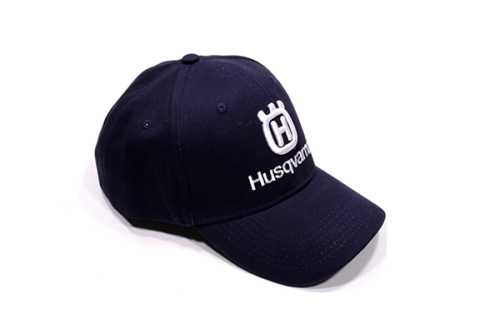 Czapka z daszkiem Husqvarna w granatowym kolorze z białym haftem logo i napisem. Pozycja produktu lekko z boku, ukazuje daszek oraz detale wykończenia. Tło jednolite, co podkreśla kształt i kolor czapki.