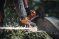Buty skórzane z ochroną przed przecięciem w kolorze pomarańczowym i czarnym, widoczne z boku na leśnym tle, stojące na pniu drzewa. Przód butów posiada wzmocnioną osłonę, a cholewka z materiału tekstylnego gwarantuje komfort i stabilność.