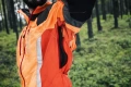 Kurtka do prac leśnych w kolorze pomarańczowym z czarnymi elementami, z widocznymi zamkami i siateczkowymi wstawkami pod pachami. Prezentowana z boku, na mężczyźnie, w leśnym otoczeniu, detale funkcyjne i ergonomiczne.