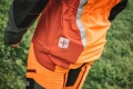 Pomarańczowo-czerwona kurtka do prac leśnych z logo na kieszeni, ukazana z boku na tle zielonej trawy. Detale materiału i kontrastowe wstawki podkreślają przeznaczenie na zewnątrz.