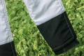 Nogawki ochronne Classic Husqvarna w kolorze białym z czarnym wykończeniem. Widoczne są detale szwów oraz mankiet. Produkt umieszczony w naturalnym otoczeniu, wśród zielonych roślin. Ujęcie z boku, ukazujące dolną część nogawki.